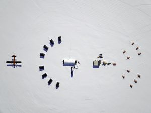 Aerial view of a snowy landscape in Antarctica with scattered tents, small buildings, snow vehicles in a semicircle, and a red plane on the left, forming an organized remote camp supported by Antarctic Logistics and Expeditions.