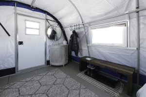 Interior of a white tent at the South Pole, featuring a door, round mirror, cabinet, coat rack with a black jacket, small window, wooden bench on a rug, and a geometric-patterned carpet—perfect for Antarctic Logistics and Expeditions.