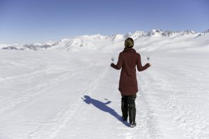 A person in a maroon coat walks on a snowy landscape in Antarctica, holding a glass in each hand, with snow-covered mountains and a clear blue sky in the background.