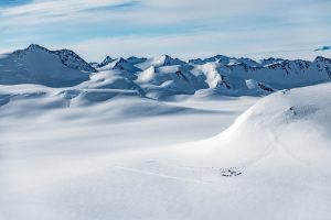 A remote camp with clustered tents sits on a vast, snowy plain in Antarctica, surrounded by rugged, snow-covered mountains under a blue sky with wispy clouds.