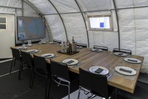 A large wooden dining table with ten black chairs is set for a meal inside a white tent in Antarctica. Bottles and an ice bucket are in the center. A map hangs on the left wall, capturing the spirit of Antarctic Logistics and Expeditions.