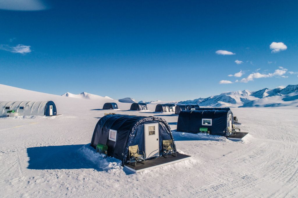 Several insulated tents with small porches are set up on a snowy, open landscape under a clear blue sky at the South Pole, with distant snow-covered mountains in the background—an expedition supported by Antarctic Logistics and Expeditions.