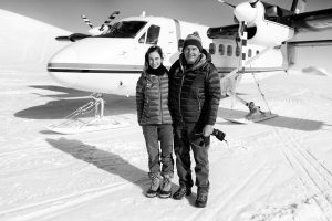 A smiling man and woman in winter clothing stand on snow beside a small propeller plane, likely with Antarctic Logistics and Expeditions. The sun casts long shadows across the remote, icy ground—perhaps near the South Pole or deep in Antarctica.
