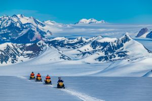 Four people ride snowmobiles across a snowy, mountainous Antarctic landscape under a clear blue sky, with rugged, snow-covered peaks visible in the background—a true adventure with Antarctic Logistics and Expeditions.