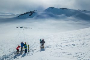 Four people with skis and snowboards stand on a snowy slope in Antarctica, preparing to descend. Vast snowfields stretch toward distant mountain peaks, with Antarctic Logistics and Expeditions tents visible on the plain below.