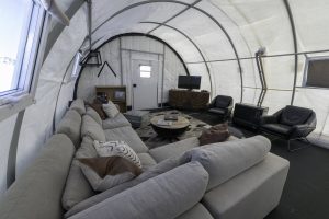 Modern living space inside a large white tent at the South Pole with a sectional sofa, armchairs, TV on a wooden stand, coffee table, and bookshelves. The room has a curved, industrial-style ceiling and windows—perfect for Antarctic Logistics and Expeditions.