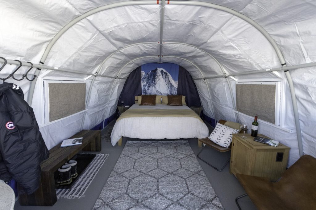 Interior of a cozy canvas tent reminiscent of Antarctic Logistics and Expeditions, featuring a bed with pillows and blankets, geometric rug, bench with boots, coat on hooks, side table with wine and glasses, and a mountain-themed wall mural behind the bed.