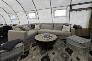 A cozy lounge area with a large beige sectional sofa, patterned throw pillows, and a round wooden coffee table invites relaxation—perfect after a day of Antarctic Logistics and Expeditions at the South Pole, inside a tent-like room with windows and shelves.