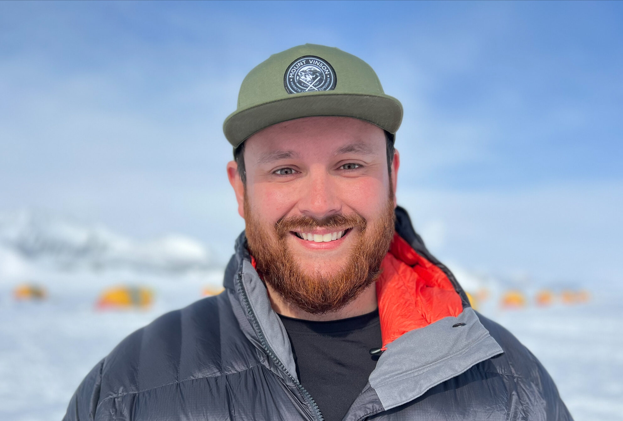 A man with a beard and green cap smiles outdoors, wearing a puffy gray jacket with a red lining. Snowy tents hint at an Antarctica adventure, with the blue sky and South Pole camp in the blurred background.