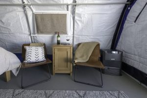 Two brown chairs with pillows and a blanket sit beside a wooden nightstand holding a wine bottle and glass, inside a cozy tent reminiscent of an Antarctic Logistics and Expeditions camp, with a bed, heater, and gray rug visible.