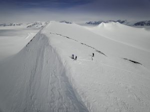 Three people hike along a narrow, snow-covered mountain ridge in Antarctica, with vast snowy plains and distant mountains under a cloudy sky—a scene reminiscent of an Antarctic Logistics and Expeditions journey.