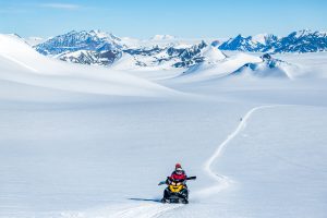 A person rides a yellow snowmobile across a vast, snowy landscape in Antarctica with distant mountains under a bright blue sky. A single snowmobile track, part of an Antarctic Logistics and Expeditions journey, winds through the untouched snow.