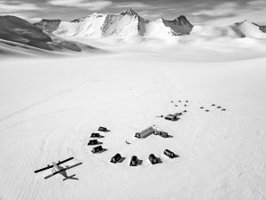 A black-and-white aerial view of an Antarctica camp shows tents in a semicircle, a small plane, and jagged peaks beyond. Tracks in the snow hint at Antarctic Logistics and Expeditions leading toward adventure near the South Pole.