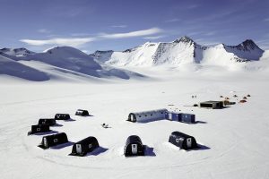 Aerial view of a snowy Antarctic landscape with several black tents and portable structures arranged in a circle, surrounded by mountains under a clear blue sky—an impressive setup by Antarctic Logistics and Expeditions near the South Pole.