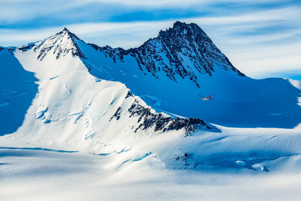 A small airplane from Antarctic Logistics and Expeditions flies over a vast, snowy mountain range with rugged peaks beneath a blue sky and wispy clouds in Antarctica.