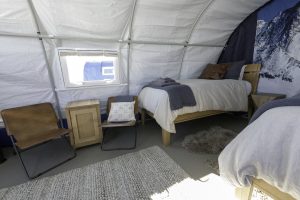 A cozy tent bedroom with two twin beds, brown leather chairs, small wooden nightstands, neutral bedding, and a fluffy rug. Natural light pours in as a mountain mural hints at the spirit of Antarctic Logistics and Expeditions in remote Antarctica.