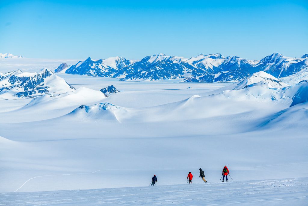 Four people in winter gear hike across a vast, snowy landscape in Antarctica, with snow-covered mountains and a clear blue sky—an epic journey reminiscent of Antarctic Logistics and Expeditions adventures to the South Pole.