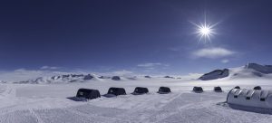 Rows of dark tents are set up on a vast, snowy landscape in Antarctica under a bright sun and clear blue sky, with distant mountains visible on the horizon.