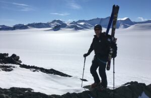 A person wearing winter gear stands on snowy rocks with skis strapped to their backpack, holding ski poles. Snow-covered mountains and a bright sky evoke the remote beauty of Antarctica, reminiscent of an Antarctic Logistics and Expeditions journey.