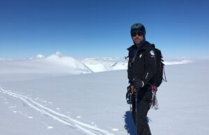 A person wearing black winter gear, helmet, and sunglasses stands on a snowy Antarctic landscape with mountains in the background under a clear blue sky, holding mountaineering equipment, ready for an Antarctic Logistics and Expeditions journey.