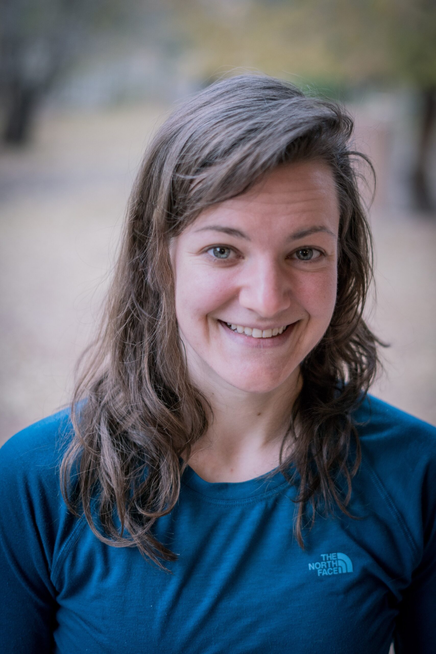 A woman with long brown hair and a blue The North Face shirt smiles at the camera outdoors, the blurred background of trees and autumn ground evoking memories of Antarctica adventures with Antarctic Logistics and Expeditions.