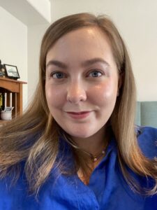 A woman with straight light brown hair and blue eyes, wearing a royal blue shirt, smiles at the camera indoors. Behind her, a wooden shelf with decorations hints at her recent return from an Antarctic Logistics and Expeditions trip.