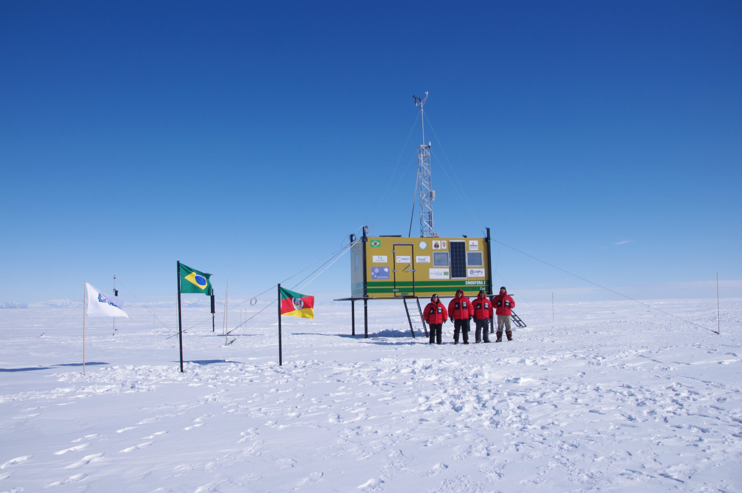 A group of five people in red jackets stands beside a yellow research station elevated on stilts in Antarctica’s snowy, flat landscape, with several flags—including Brazil’s—flying nearby under the clear South Pole sky.