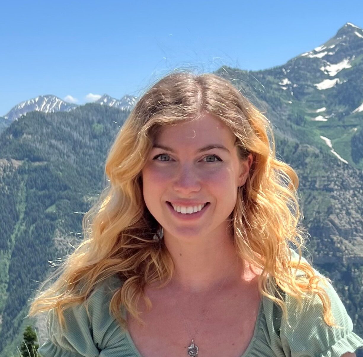 A young woman with wavy blonde hair, wearing a light green top and a necklace, smiles outdoors with snow-capped mountains and green hills in the background under a clear blue sky, capturing the spirit of Antarctic Logistics and Expeditions.