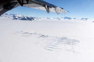 Aerial view of a snowy, remote scientific outpost in Antarctica, supported by Antarctic Logistics and Expeditions, with small buildings and equipment amid vast ice and mountain ranges under a clear blue sky. Part of an aircraft wing is visible overhead.