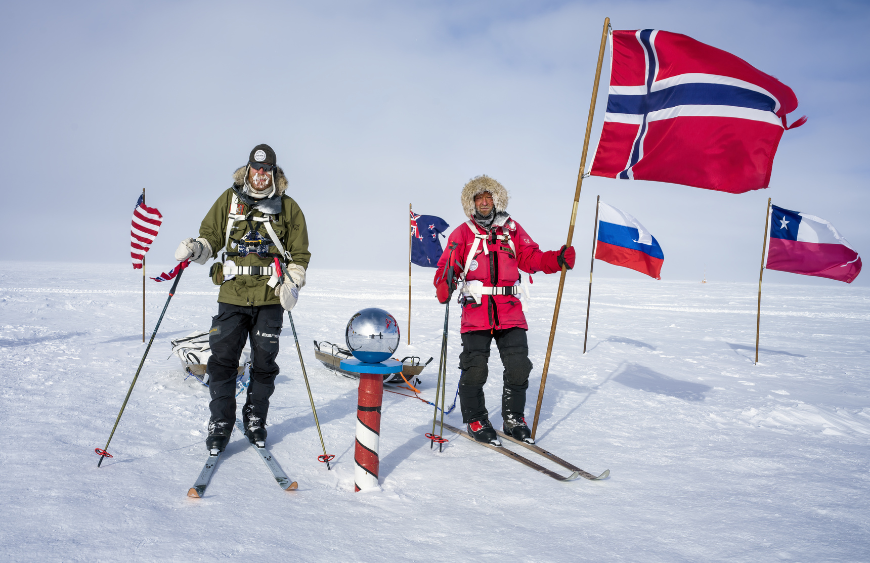 Two people in cold-weather gear stand on snow near the South Pole marker in Antarctica, holding ski poles. Large flags of Norway, the USA, and others wave in the wind—a scene made possible by Antarctic Logistics and Expeditions.