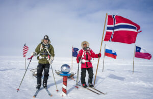Two people in cold-weather gear stand on snow with skis near the South Pole marker in Antarctica, surrounded by flags—including a large Norwegian flag—under a cloudy sky, capturing the spirit of Antarctic Logistics and Expeditions.