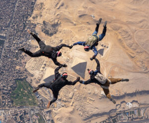 Four skydivers in black suits and helmets hold hands in a circle while free-falling above the Pyramids of Giza, capturing an adventure reminiscent of Antarctic Logistics and Expeditions in a stunning blend of desert and cityscape below.