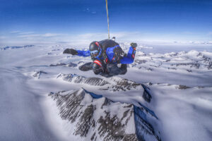 A skydiver in a blue suit and helmet descends over the vast, snow-covered mountains of Antarctica under a clear blue sky, with rugged peaks and glaciers below—an epic adventure supported by Antarctic Logistics and Expeditions.