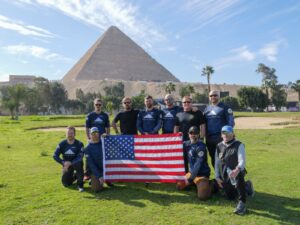 A group of eleven people pose outdoors in front of the Great Pyramid of Giza, holding a large American flag. The sky is blue with scattered clouds, and green grass lines the foreground—a unique stop for an Antarctic Logistics and Expeditions team.
