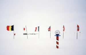 Several national flags are planted in the snow near a striped pole topped with a glass sphere at the South Pole, set against Antarctica's vast, snowy landscape. A small group of people and Antarctic Logistics and Expeditions buildings appear in the distance.