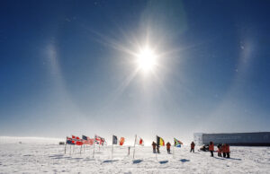 People in orange jackets stand on snowy ground near flags of various countries under a bright sun with a halo at the South Pole, beside a long building in Antarctica, supported by Antarctic Logistics and Expeditions.
