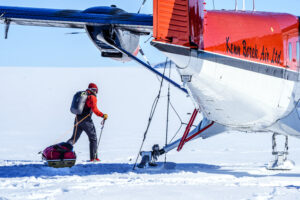 A person in cold-weather gear pulls a sled across snow toward a red and white plane labeled Kenn Borek Air Ltd, part of Antarctic Logistics and Expeditions, on an icy, remote landscape in Antarctica.