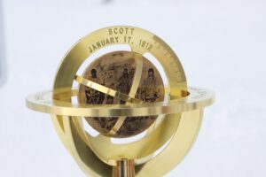 A close-up of a brass sundial featuring an engraved globe with explorers, inscribed Scott January 17, 1912. The snowy, out-of-focus background evokes the harsh beauty of Antarctica and the spirit of South Pole exploration.