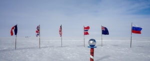 Six international flags stand in the snow near a striped pole topped with a shiny silver sphere, marking the South Pole against Antarctica’s vast, snowy, and clouded landscape.
