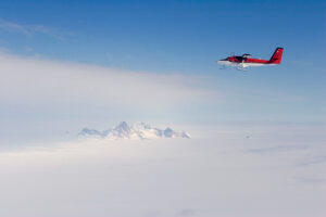A red and white small airplane flies above a vast, snowy Antarctic landscape with a mountain peak visible through the clouds below. Operated by Antarctic Logistics and Expeditions, it soars beneath a mostly clear, blue sky.