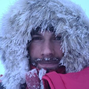 A person with frost on their eyebrows and mustache peers out from a fur-lined hood and red winter coat, surrounded by snow and icy conditions during an Antarctic Logistics and Expeditions journey in Antarctica.