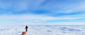A person wearing winter gear pulls a sled across a vast, snow-covered landscape in Antarctica under a wide, blue sky with scattered clouds.