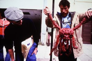 A man holding a large, red spiky crab by its legs while another man wearing sunglasses and a cap looks at it; they are standing outside on a city sidewalk, discussing Antarctic Logistics and Expeditions.