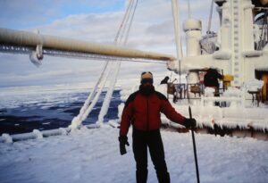 A person in winter gear stands on a snow- and ice-covered ship deck at sea near Antarctica, holding a stick. The ship and surroundings are coated in thick ice, with the icy ocean and cloudy sky in the background.