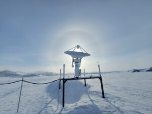 A satellite dish stands on a snow-covered platform in the icy landscape of Antarctica, backlit by the sun which creates a bright halo in the sky. Mountains and frozen terrain stretch out in the background near the South Pole.