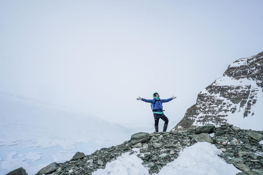 A person in winter gear stands on a snowy, rocky slope with arms outstretched. Snow blankets the ground near Antarctica’s vast peaks, while the pale, overcast sky hints at the remote adventure toward the South Pole.