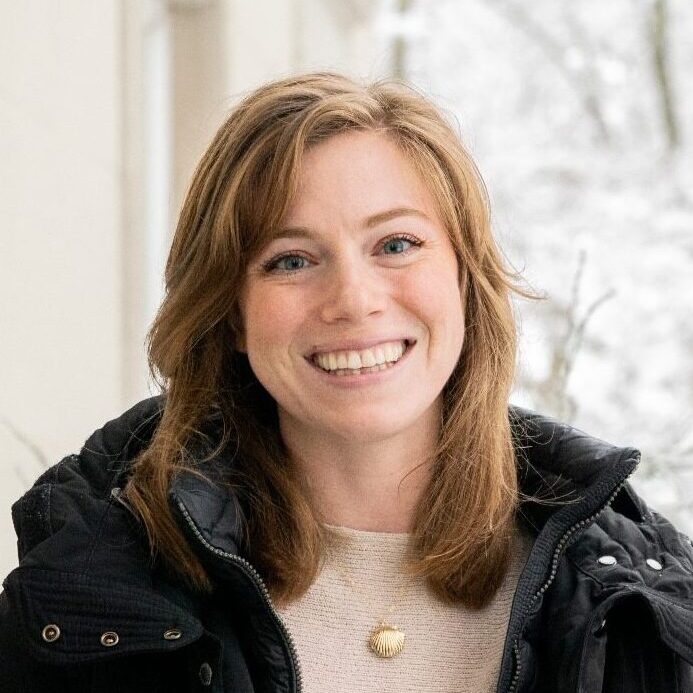A woman with shoulder-length light brown hair smiles at the camera. Wearing a black jacket over a light sweater and a round pendant necklace, she stands before a softly blurred, wintery scene reminiscent of Antarctica.