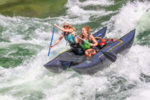 Two women paddle a blue inflatable kayak through whitewater rapids, smiling and excited. Both wear life jackets; one has a hat and sunglasses. Water splashes as they navigate the rough river, reminiscent of an Antarctic Logistics and Expeditions adventure.