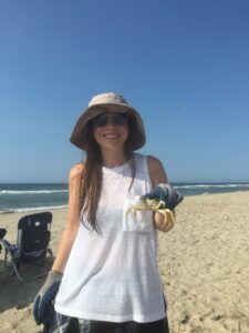A smiling woman wearing a sunhat, sunglasses, gloves, and a white shirt holds a crab on a sandy beach, with waves and a blue sky in the background. She dreams of Antarctic Logistics and Expeditions as people relax by the shore nearby.