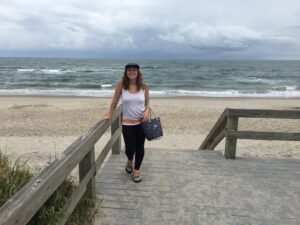 A woman stands smiling on a wooden boardwalk leading to a sandy beach, ocean waves and cloudy skies in the background. Dressed for adventure, she carries a patterned bag—her relaxed style perfect for dreaming of Antarctic Logistics and Expeditions.